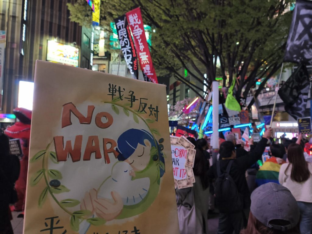 写真。デモの様子を背景に、Tokinのイラストのプラカード。戦争反対、NO WAR と書いてある。