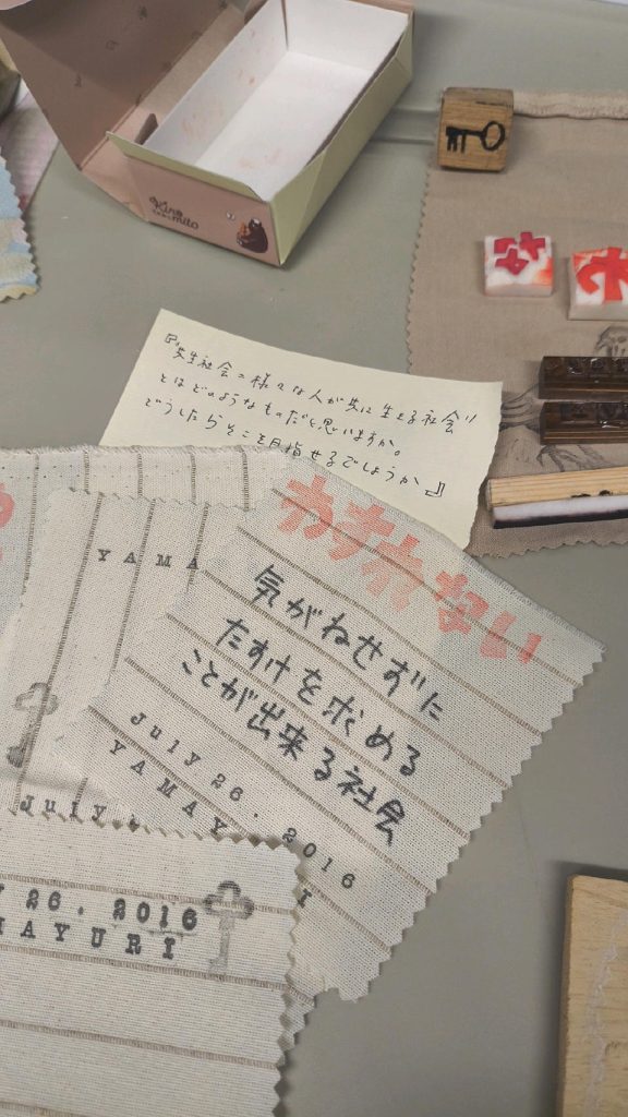 スタンプなどが置いてあるテーブル上の写真。「共生社会=さまざまな人が共に生きる社会、とはどのようなものだと思いますか」というメモの下に。「わすれない」というスタンプがおしてある布。マジックで「気兼ねせずに助けを求めることができる社会」と書いてある。